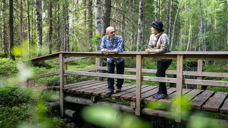 Kaksi ihmista nojailee puusillan kaiteeseen metsäpurolla.