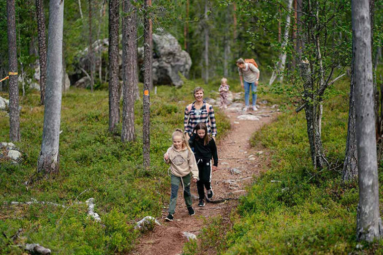 Kaksi aikuista ja kolme lasta kävelevät polulla metsässä. Taustalla on suuri kivenlohkare.