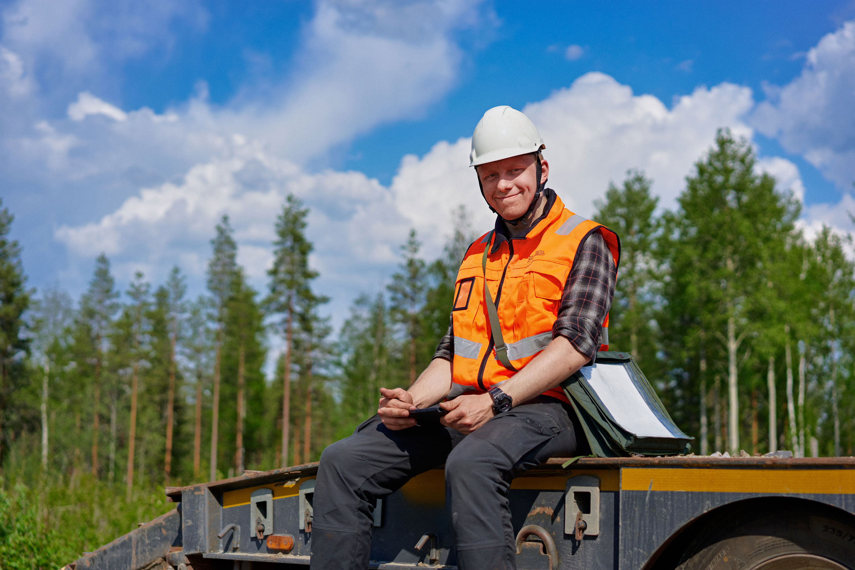 Oranssiliivinen metsätalouden toimihenkilö kypärä päässä istumassa lavetin päällä.