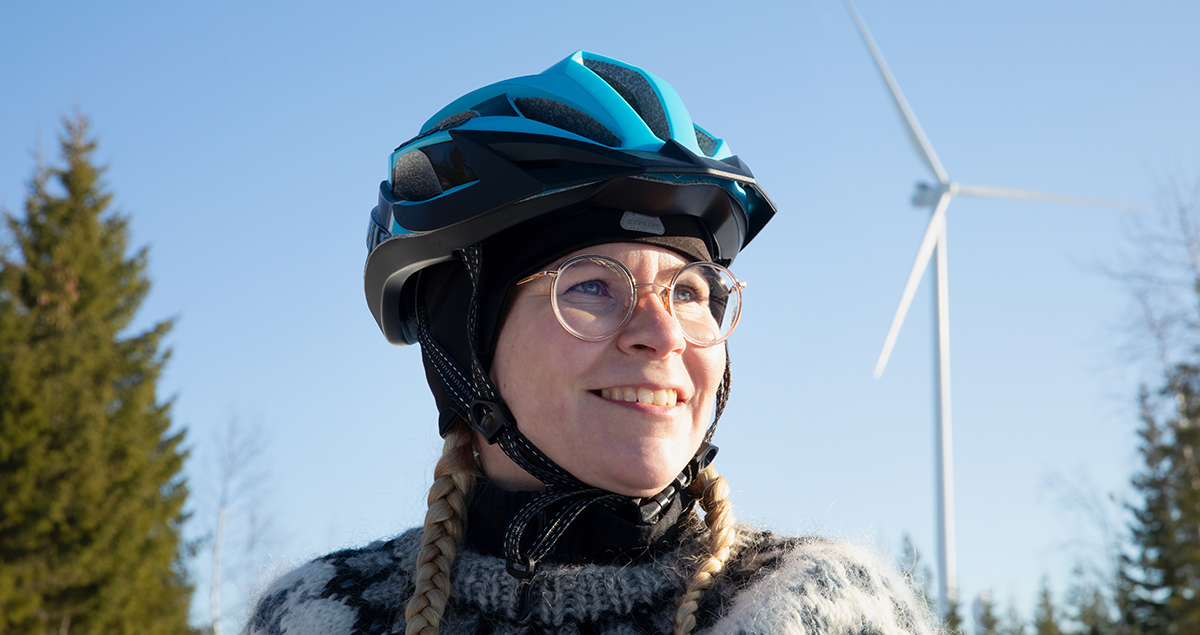 En kvinna i närbild med cykelhjälm och vindkraftsmast syns bakom.