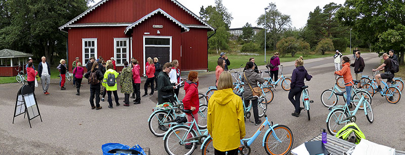 Bakom finns ett rött trähus. Framför är ett torg med många människor. Vissa har cyklar, andra inte. Människorna väntar på att guidningen ska börja.