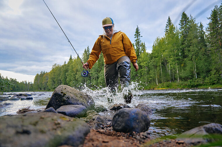 Perhokalastaja astumassa vedestä rannalle vapa kädessään.