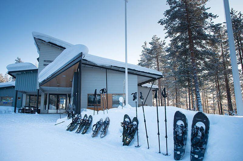 På taket av den ljusa träbyggnaden ligger snö. På gården står snöskor och stavar i rad i snön.