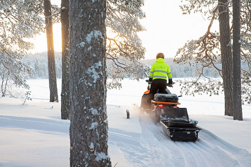 Metsähallituksen työntekijä ajaa moottorikelkalla lumisessa maastossa. Aurinko paistaa matalalta.